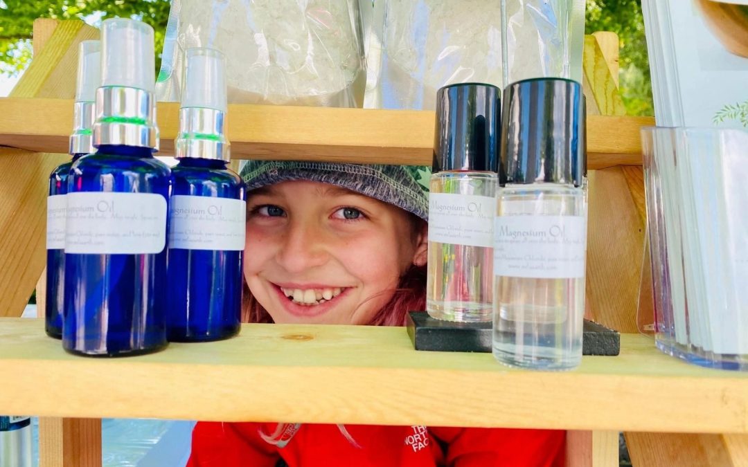 Smiling child peeking out from behind a shelf of Mila Earth magnesium bottles at a market stall