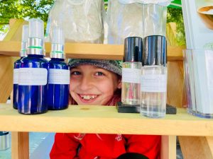 Smiling child peeking out from behind a shelf of Mila Earth magnesium bottles at a market stall
